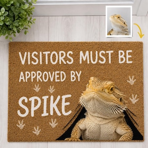 Può includere: Uno zerbino rettangolare in fibra di cocco marrone con il testo bianco "VISITORS MUST BE APPROVED BY SPIKE", un'immagine stampata di un drago barbuto e diverse icone di impronte di lucertola bianche sulla superficie ruvida.