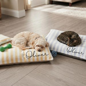 May include: Two rectangular pet beds with the name Oliver embroidered in black script. One bed features yellow and white vertical stripes with a small green toy, while the other has blue and white vertical stripes. A dog and a cat are sleeping.