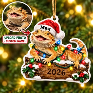 May include: A custom Christmas ornament featuring a bearded dragon wearing a Santa hat with the name Max printed on it. The lizard rests on a snowy log decorated with colourful string lights and holly, with the year 2026 printed on the wood surface.