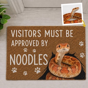 May include: A rectangular brown coir doormat featuring the white text VISITORS MUST BE APPROVED BY NOODLES. The mat displays a graphic of a coiled orange and brown snake and several white paw prints. A small inset image shows the snake.