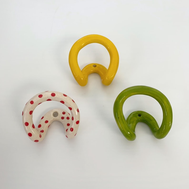 May include: Three ceramic wall hooks with a curved, rounded shape. One hook is solid yellow, one is solid lime green, and one is off-white with small red polka dots. Each hook features a central hole for mounting, showcasing a smooth, glossy finish.