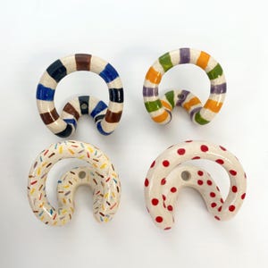 Könnte beinhalten: Vier Wandhaken aus Keramik mit geschwungenem, kreisförmigem Design. Ein Haken hat blaue und braune Streifen, ein weiterer grüne, orange und violette Streifen, einer ein buntes Streuselmuster und der letzte rote Punkte auf cremefarbenem Grund.