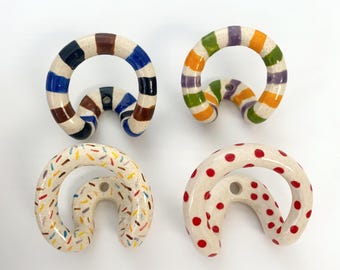 May include: Four ceramic wall hooks with a curved, circular design. One hook features blue and brown stripes, another has green, orange, and purple stripes, one has a multicolored sprinkle pattern, and the last has red polka dots on a cream base.