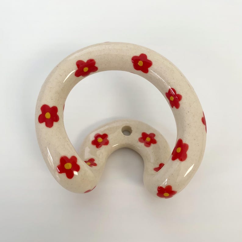 May include: A circular wall hook with a speckled off-white finish, decorated with small red flowers featuring yellow centers. The hook has a curved, U-shaped base with a single mounting hole at the top center for attachment to a wall surface.