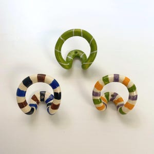 May include: Three ceramic wall hooks with a curved, circular design. One hook is solid lime green with thin white stripes. The other two hooks feature a speckled cream base with alternating horizontal stripes in blue, brown, black, orange, and purple.