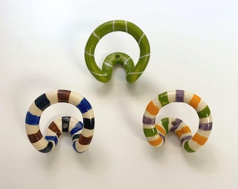 May include: Three ceramic wall hooks with a curved, circular design. One hook is solid lime green with thin white stripes. The other two hooks feature a speckled cream base with alternating horizontal stripes in blue, brown, black, orange, and purple.