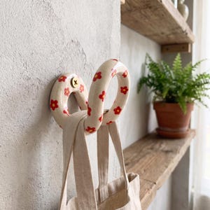 May include: A cream-colored ceramic wall hook with a rounded, looped shape featuring a repeating pattern of small red flowers with yellow centers. The hook is mounted to a textured wall with a single gold-toned screw and holds a beige fabric bag.
