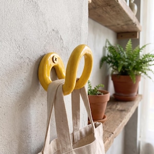 May include: A bright yellow, curved ceramic wall hook mounted with a single screw, featuring a smooth, glossy finish. A beige canvas tote bag with two fabric handles is hanging from the hook, which is attached to a textured gray wall surface.