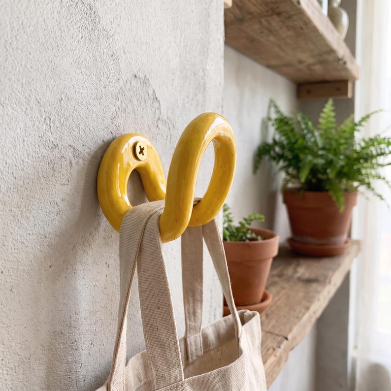 May include: A bright yellow, curved ceramic wall hook mounted with a single screw, featuring a smooth, glossy finish. A beige canvas tote bag with two fabric handles is hanging from the hook, which is attached to a textured gray wall surface.