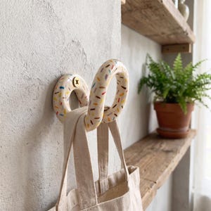 May include: A wall-mounted ceramic hook shaped like a donut with a cream-colored glaze and colorful sprinkles. The hook is secured with a brass screw and holds a plain beige canvas tote bag with two fabric handles hanging from its curved form.
