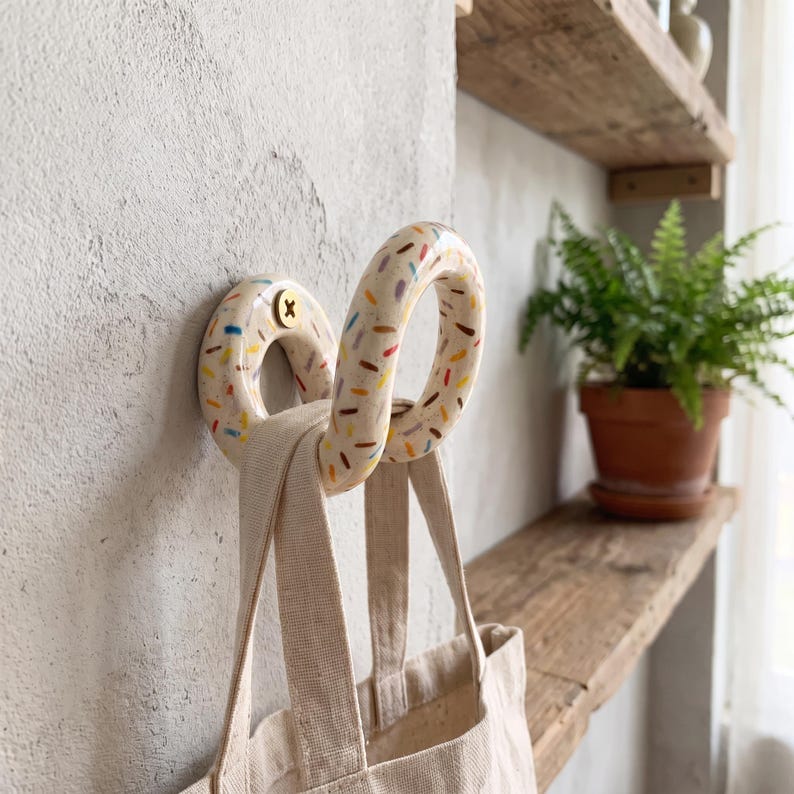 May include: A wall-mounted ceramic hook shaped like a donut with a cream-colored glaze and colorful sprinkles. The hook is secured with a brass screw and holds a plain beige canvas tote bag with two fabric handles hanging from its curved form.