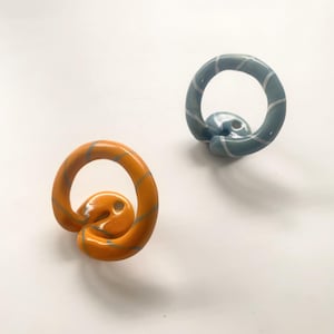 Könnte beinhalten: Zwei Wandhaken aus Keramik mit glänzender Oberfläche. Ein Haken ist leuchtend orange mit dünnen, hellgrauen diagonalen Streifen, der andere ist blaugrau mit ähnlichen hellgrauen Streifen. Jeder Haken hat ein kreisförmiges, geschlungenes Design.