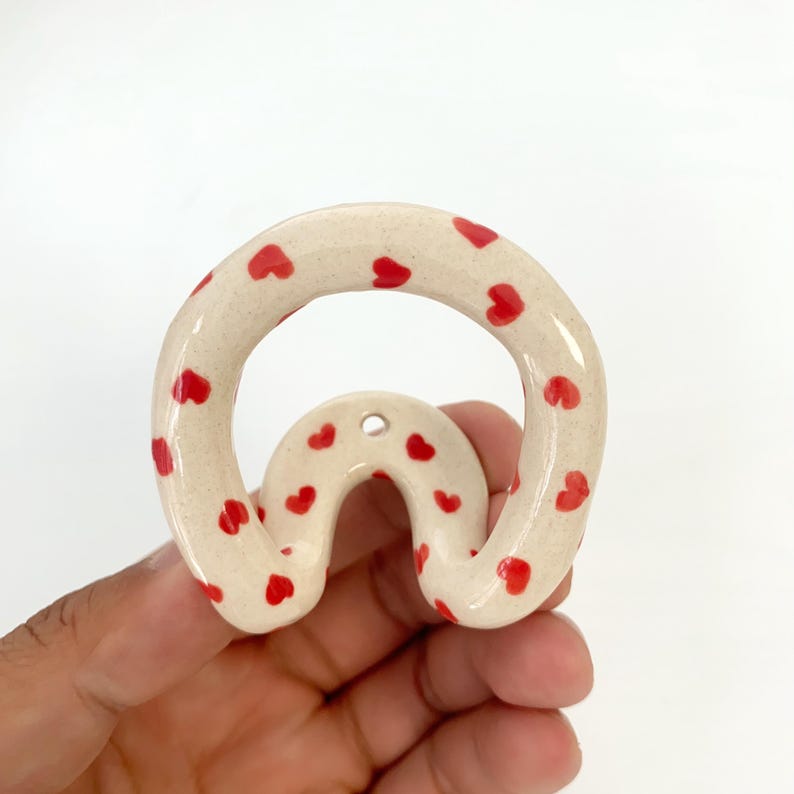 May include: A cream-colored ceramic wall hook with a speckled texture and a repeating pattern of small red hearts. The hook features a rounded, arch-like shape with a single mounting hole at the top center, designed for hanging items on a wall.