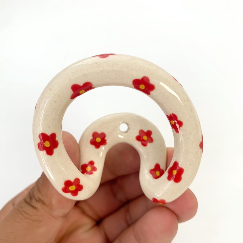 May include: A cream-colored ceramic wall hook with a curved, arch-like shape. The surface features a speckled texture and is decorated with multiple small, hand-painted red flowers with yellow centers. A single mounting hole is visible at the top.