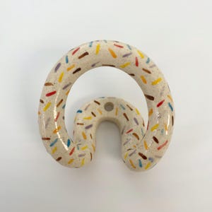 May include: A ceramic wall hook with a speckled cream-colored glaze and a pattern of small, multi-colored dash-like markings in red, yellow, blue, and brown. The hook has a curved, rounded shape with a single mounting hole at the top center.