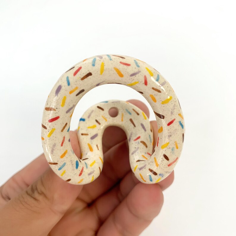 May include: A ceramic wall hook with a curved, arch-like shape and a speckled cream-colored glaze. The surface is decorated with small, scattered dash-shaped patterns in various colors including red, yellow, blue, brown, and purple.
