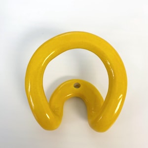 May include: A bright yellow wall hook with a smooth, glossy finish. The hook features a circular, open-loop design that curves downward, with a single small circular mounting hole positioned at the top center of the curved structure.