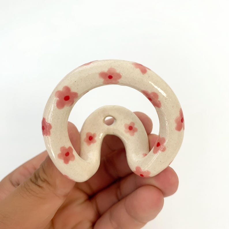 May include: A cream-colored ceramic wall hook with a rounded, arch-like shape. The surface is decorated with small, hand-painted pink flowers featuring red centers. A single circular mounting hole is positioned at the top of the arch structure.