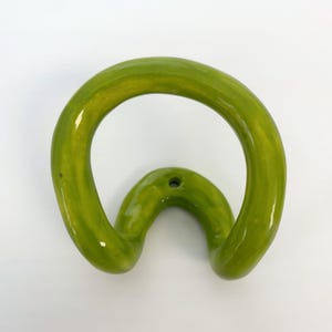 May include: A lime green ceramic wall hook with a glossy finish. The hook features a continuous, rounded, organic loop shape that curves outward from a central mounting point, which includes a single small hole for installation on a wall surface.