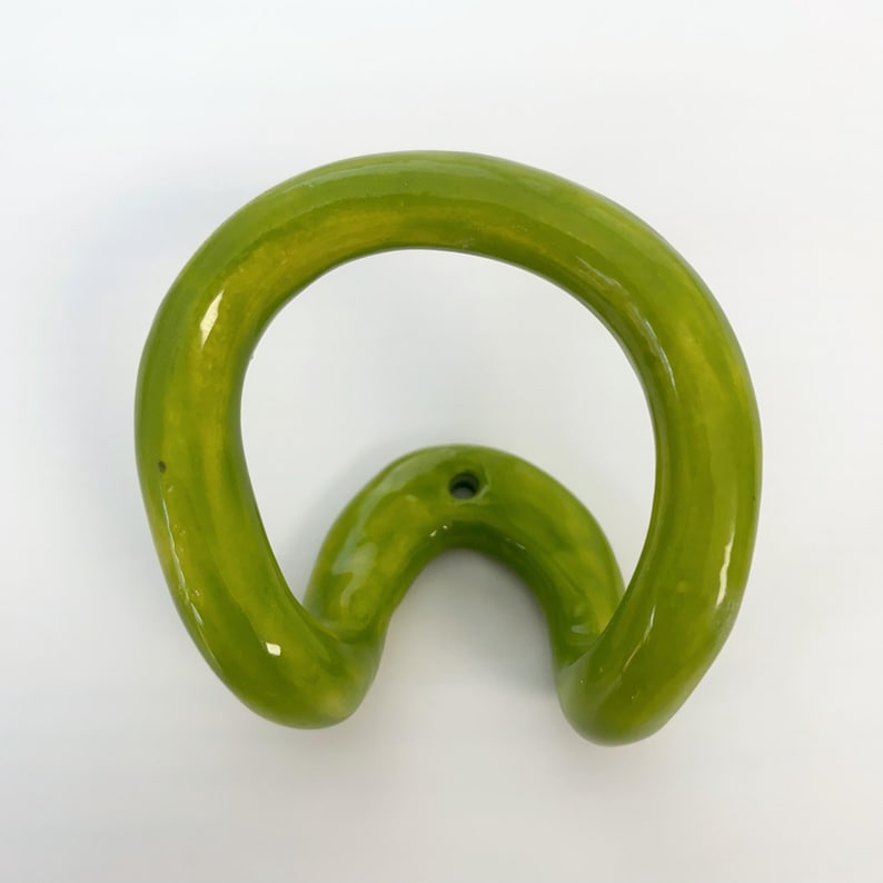 May include: A lime green ceramic wall hook with a glossy finish. The hook features a continuous, rounded, organic loop shape that curves outward from a central mounting point, which includes a single small hole for installation on a wall surface.