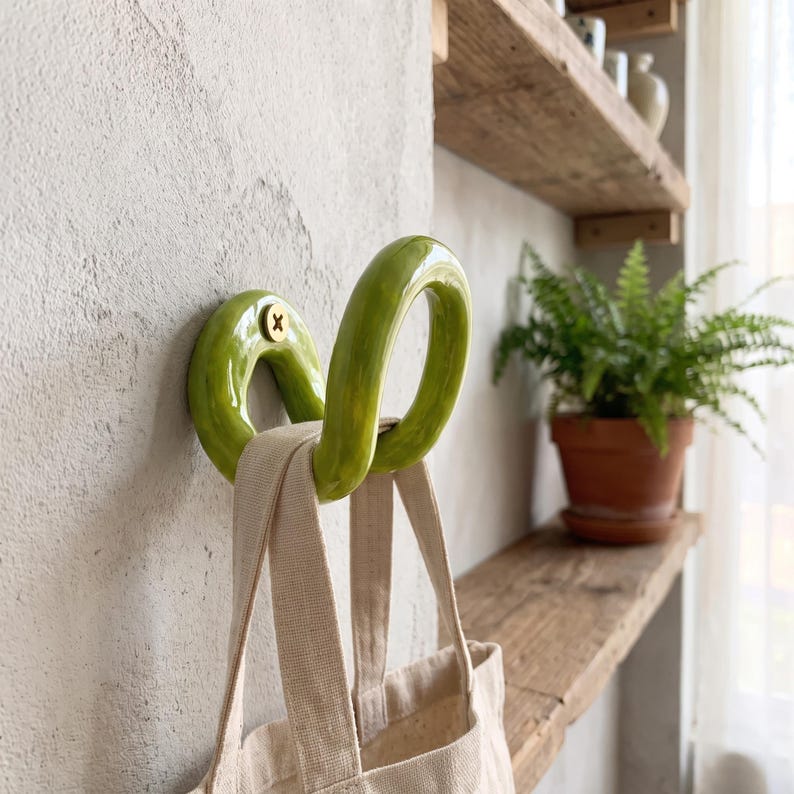 May include: A lime green ceramic wall hook with a curved, looped design mounted to a textured wall with a gold-colored screw. A beige canvas tote bag with two fabric handles hangs from the hook, which features a glossy, smooth finish.