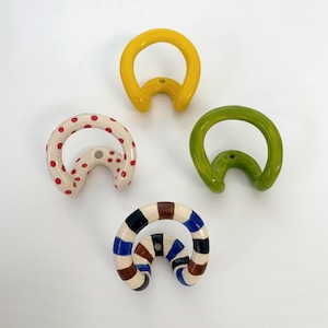 Op de afbeelding: Vier wandhaken met een gebogen, cirkelvormig ontwerp. Eén is effen geel, één is effen groen, één is crème met rode stippen en één is crème met blauwe, bruine en zwarte horizontale strepen. Elke haak is voorzien van een centraal montagegat.