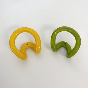 May include: Two wall hooks with a glossy finish, one in bright yellow and one in lime green. Each hook features a curved, continuous loop design with a single circular mounting hole at the base for attachment to a flat surface.