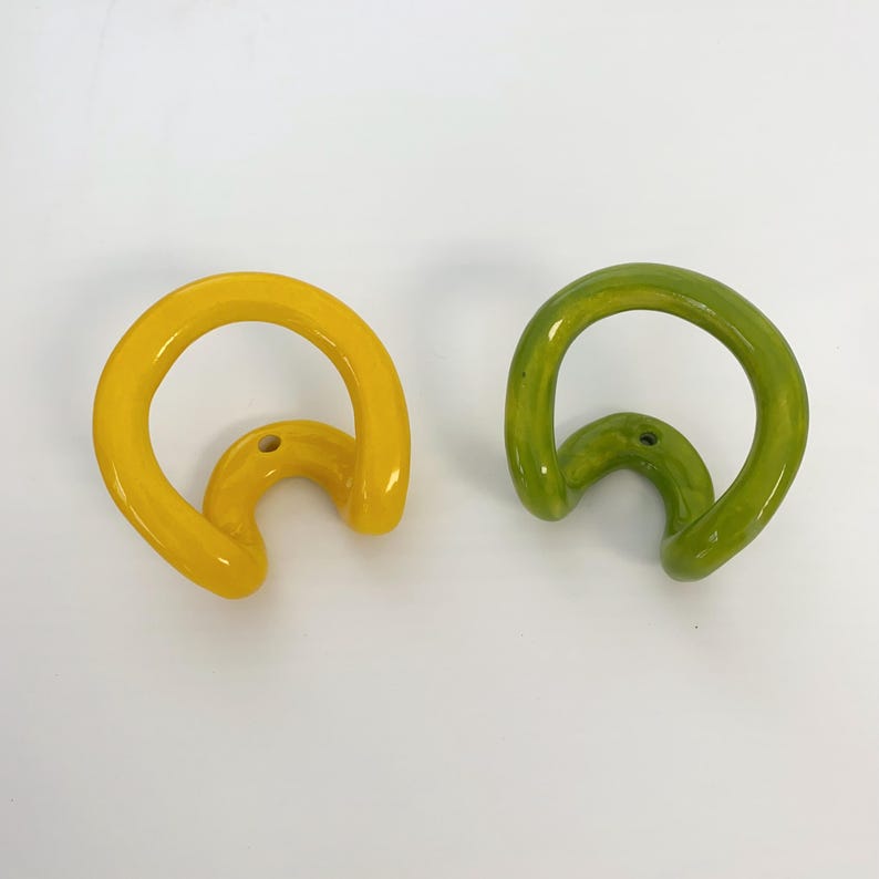 May include: Two wall hooks with a glossy finish, one in bright yellow and one in lime green. Each hook features a curved, continuous loop design with a single circular mounting hole at the base for attachment to a flat surface.