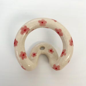 May include: A cream-colored ceramic wall hook with a circular front loop and a curved base featuring a single mounting hole. The surface is decorated with hand-painted light pink flowers, each accented with a small, vibrant red dot in the center.
