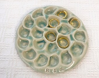 May include: A round, light blue and white glazed ceramic ring dish with a textured, honeycomb-like surface featuring multiple indentations. The dish has the letters R & A stamped near the bottom edge and holds four gold-toned metal rings.