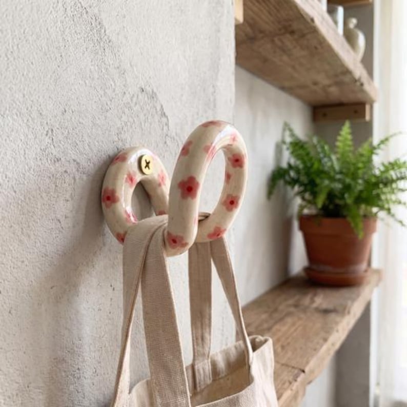 May include: A cream-colored ceramic wall hook with a hand-painted pink flower pattern. The hook has a rounded, looped shape and is secured to the wall with a single brass-colored screw. A beige canvas tote bag hangs from the ceramic hook.