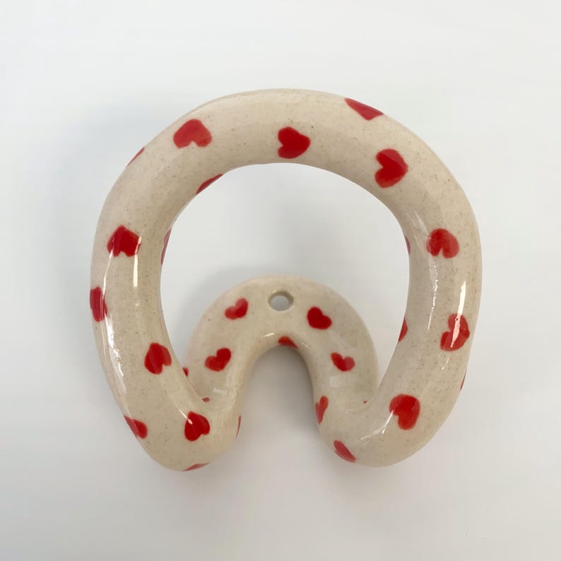 May include: A ceramic wall hook with a glossy, speckled off-white finish and a pattern of small, hand-painted red hearts. The hook features a curved, organic shape with a single circular mounting hole at the top center for attachment to a wall.