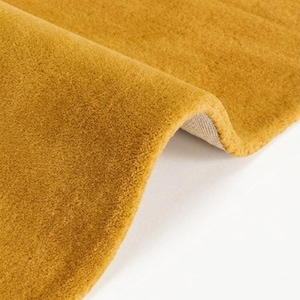 Könnte beinhalten: Ein einfarbiger senfgelber Teppich mit dichter Kurzflor-Struktur. Der Teppich ist umgeschlagen und gibt den Blick auf eine hellbeige, gewebte Stoffrückseite frei, die die Dicke und die Beschaffenheit des textilen Materials verdeutlicht.