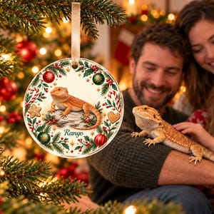 May include: A round, flat, clear acrylic Christmas ornament featuring a printed illustration of a bearded dragon lizard resting on a rock. The design is framed by a wreath of green pine needles, red berries, candy canes, and ornaments with the name Rango.