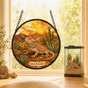 Peut inclure: Un attrape-soleil circulaire en vitrail représentant un dragon barbu dans un paysage désertique avec un cactus, des plantes grasses et un coucher de soleil. Le nom RANGO est imprimé en lettres noires sur une bannière. Chaîne métallique noire.