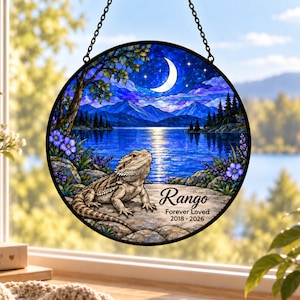 May include: A circular stained glass suncatcher featuring a bearded dragon resting on a rock by a lake at night. The scene includes mountains, a crescent moon, and stars. The text reads Rango Forever Loved 2018 - 2026 in black script and print.
