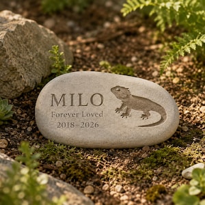 May include: A smooth, light grey oval memorial stone featuring an engraved image of a bearded dragon. The text "MILO", "Forever Loved", and the years "2018-2026" are etched into the surface of the stone in a clear, dark, serif font.