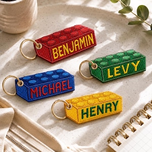 May include: Four rectangular embroidered keychains shaped like building blocks in red, green, blue, and yellow. Each features a gold-tone metal ring and is personalized with stitched names: BENJAMIN, LEVY, MICHAEL, and HENRY in contrasting thread.
