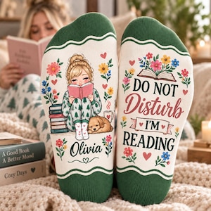 May include: A pair of cream-colored socks with dark green cuffs. The left sock features a blonde character reading a book next to a dog and a stack of books with the name Olivia. The right sock displays the text DO NOT DISTURB I'M READING.