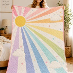 May include: A quilted blanket featuring a sun design with radiating pastel-coloured rays in pink, yellow, blue, and green. The quilt includes white clouds and small scattered stars across the surface, finished with a pink fabric border trim.