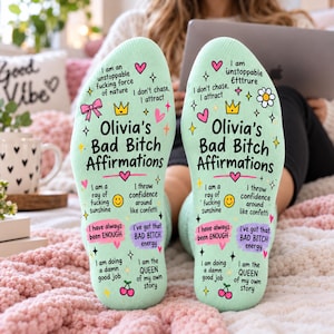 May include: A pair of light mint green socks featuring black text that reads Olivia's Bad Bitch Affirmations, surrounded by colorful icons including hearts, stars, a crown, a bow, a smiley face, and cherries, along with various motivational phrases.