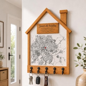 Può includere: Un organizer da parete in legno a forma di casa con cinque ganci in metallo nero. Presenta una stampa di una mappa con un cuore rosso e una targa in legno incisa con "James & Sophia, The Grand Hotel, 52 95 40 N, 1 15 50 W, 25th August 2026".