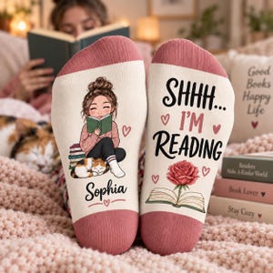 以下が含まれることがあります： ピンクの袖口が付いたクリーム色の靴下。左側には本を読む人物と猫、Sophiaという名前のイラストが描かれています。右側にはSHHH... I'M READINGという文字と、バラの花、開いた本のイラストがデザインされています。
