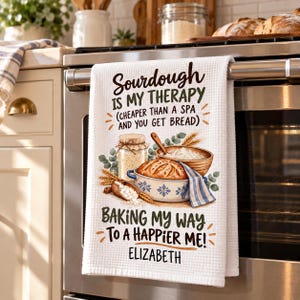 May include: A white waffle-weave kitchen towel featuring the text Sourdough is my therapy, cheaper than a spa and you get bread, and Baking my way to a happier me! Elizabeth. The design includes illustrations of a sourdough loaf, flour, and wheat.