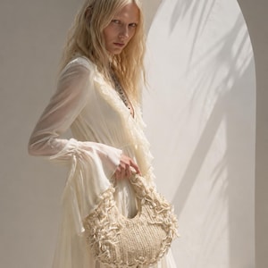 May include: A cream-colored, crescent-shaped handbag featuring a textured, woven knit construction. The exterior is embellished with numerous small, white seashells attached along the edges and across the front surface of the bag.