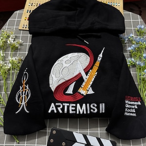Op de afbeelding: Een zwarte hoodie met een geborduurde Artemis II missiepatch op de borst, met een maan, een raket en een rode boog. De tekst ARTEMIS II staat eronder. De mouwen zijn voorzien van een raketafbeelding en de namen van de bemanningsleden.