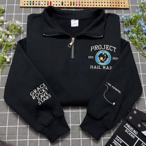 Könnte beinhalten: Ein schwarzes Sweatshirt mit Viertelreißverschluss und einer weißen Stickerei auf der Brust mit der Aufschrift PROJECT HAIL MARY EST. 2021. Der linke Ärmel zeigt den Text GRACE ROCKY SAVE STARS, der rechte Ärmel den Text I HAD POTENTIAL.