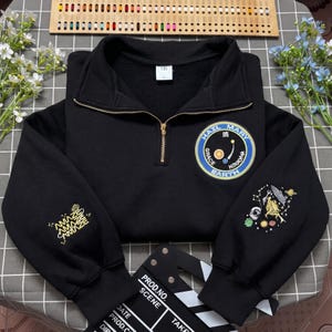Puede incluir: Una sudadera negra con cremallera corta que presenta un parche circular en el pecho con el texto Hail Mary Grace Earth y gráficos celestiales. Las mangas muestran diseños bordados que incluyen la palabra Amaze y un pájaro estilizado.