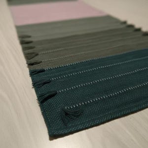 May include: A rectangular table runner featuring a woven texture with a color-blocked gradient pattern. The design transitions through shades of deep teal, forest green, olive, and a soft pale pink, finished with small decorative fringe tassels.