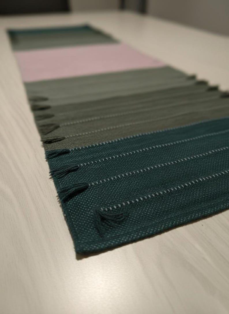 May include: A rectangular table runner featuring a woven texture with a color-blocked gradient pattern. The design transitions through shades of deep teal, forest green, olive, and a soft pale pink, finished with small decorative fringe tassels.
